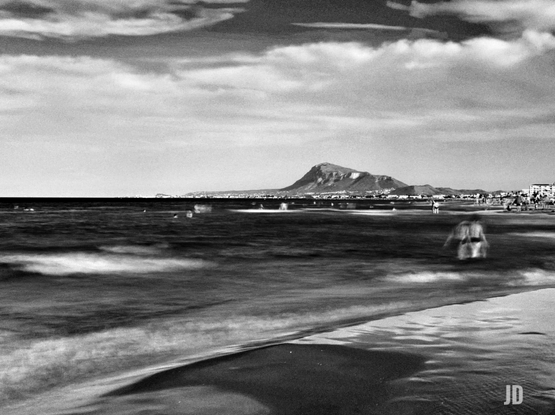 Fotografía en blanco y negro de una playa con el mar en primer plano. El cielo está parcialmente cubierto de nubes. En el horizonte, una gran montaña rocosa domina el paisaje, con una ciudad costera extendiéndose a sus pies. El mar presenta olas suaves y difuminadas, efecto de una larga exposición, con varias figuras humanas borrosas, algunas dentro del agua y otras en la orilla. La arena oscura de la playa ocupa la parte inferior derecha de la imagen.