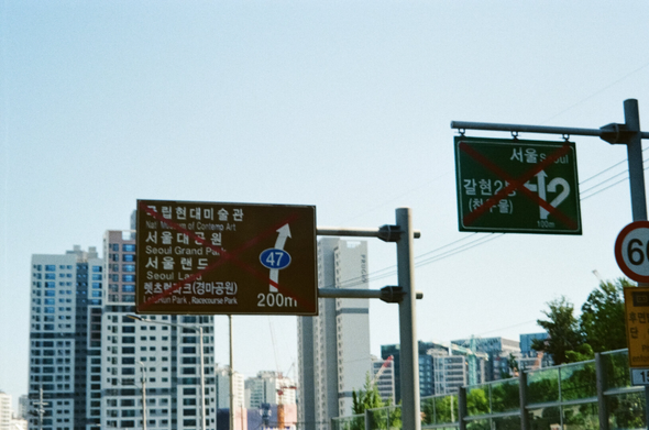 서울로 향하던 “새, 사람 행진” 도중 찍은 두 표지판. 모두 빨간색 X자가 크게 그어져 더는 사용하지 않는다는 표시가 되어있다. 왼쪽 갈색 표지판에는 “국립현대미술관·서울대공원·서울랜드·렛츠런파크(경마공원)”이, 왼쪽 초록 표지판에는 “서울”, “갈현2동(찬우물)”로 가는 표지가 그려져있다. 멀리 아파트 단지가 보인다. 카메라 고장으로 초점이 잘 맞지 않았다.