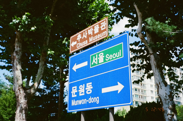 남태령 고개를 넘기 전 마주친 표지판. 거대한 파란색 표지판에 “좌측 '서울', 우측 '문원동'”이라고 써있고, 그 위에 작은 갈색 표지판에 “우측 '추사 박물관'”이라고 써있다. 뒤쪽으로 빽빽한 가로수가 있고 그 뒤로 얼핏 아파트가 보인다. 카메라 고장으로 초점이 잘 맞지 않았다.