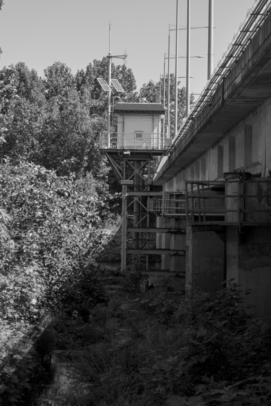 A black and white image of a steel and concrete bridge adjacent a green park with foliage, separated by a utility platform