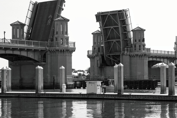 Black and white image of a drawbridge raised to allow boats to pass. The calm water reflects the bridge's structure, creating a serene atmosphere.