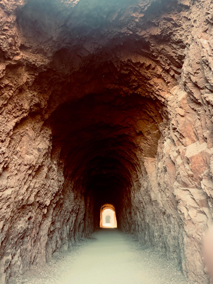 A tunnel with textured, rocky walls, leading to a brightly lit opening at the far end. The ground is gravelly, and the lighting creates a warm ambiance inside the tunnel.