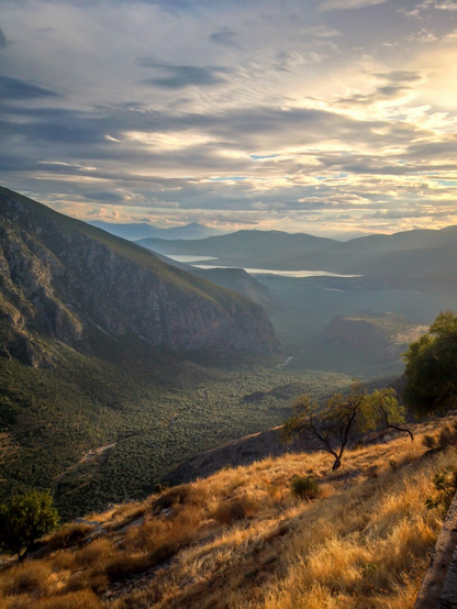 A sweeping mountain landscape at sunset, with rugged cliffs on the left, rolling hills fading into misty layers in the distance, and golden light illuminating the dry grasses and scattered trees in the foreground. The sky is filled with dramatic clouds glowing with soft tones of blue, gray, and gold.