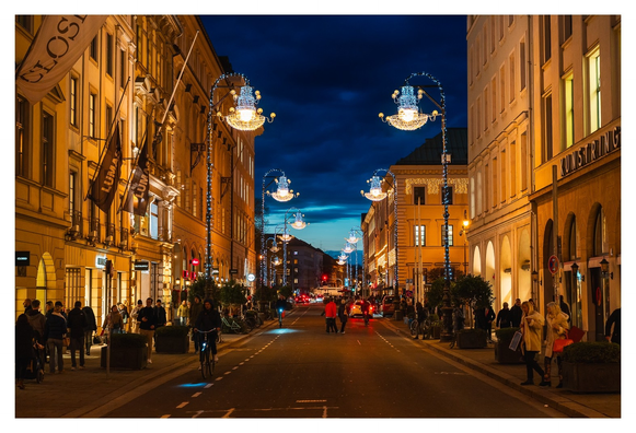 Breite Straße gesäumt von hohen Gebäuden, erleuchtet von weihnachtlich dekorierten Straßenlaternen und anderem Lichtergelump.