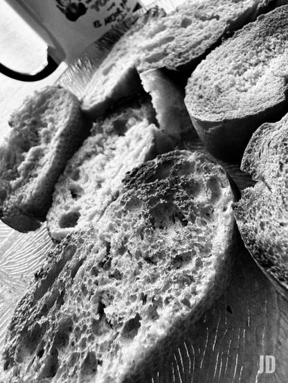 Una fotografía en blanco y negro muestra un primer plano de varias rebanadas de pan rústico o baguette. Las rebanadas tienen una miga muy aireada y porosa, llena de agujeros irregulares de diferentes tamaños, y algunas partes de la corteza, más oscuras, sugieren que el pan está tostado o bien horneado. Las rebanadas están dispuestas de forma dispersa, llenando la mayor parte del encuadre y vistas desde un ángulo ligeramente elevado. En la esquina superior izquierda, se vislumbra el borde de una taza o jarra blanca con texto borroso. El pan reposa sobre una superficie con un patrón de líneas finas y paralelas, posiblemente una tabla o un mantel texturizado. La iluminación fuerte acentúa las texturas y los contrastes de claroscuro.