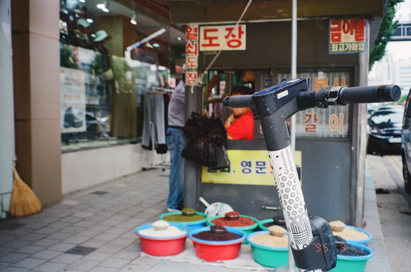 전통시장 옆 인도에 설치된 노점상에 빨간 옷을 입은 주인이 창밖을 내다보고 있다. '도장', '열쇠' 간판이 붙은 노점상 옆으로 대야에 여러 곡물류를 담아 내놓았다. 카메라 앞에 있는 킥보드 손잡이에 주인 얼굴이 가려졌다. 초점은 킥보드에 맞았다.
