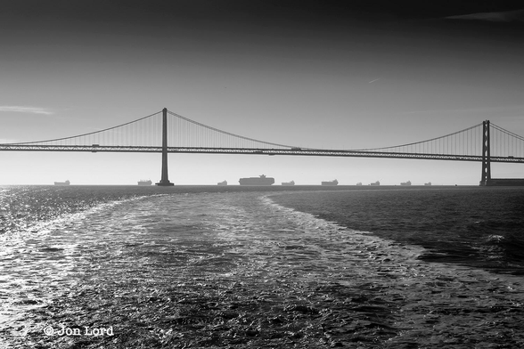 This is a black and white seascape photo of a group of ships at anchor beyond a suspension bridge. 
In the foreground is a body of dark water interrupted by speckles of white created by a ship's wake. This water rises to a little less than halfway  upwards in the image. Here there is a crisp and defined horizon with ten large cargo ship at anchor and aligned at right angles to the camera with their bows facing to the left. Between these vessels and about one km from the viewer is a large and long suspension bridge. Two towers and the deck between are in view with a further length of deck stretching out of view to the left. The bridge has two levels with lorry traffic clearly visible driving on both the upper and lower decks. Above is a near clear and cloudless sky graduating from white close to the horizon to grey/black at the top of the image. 
The location: San Francisco Bay, California. The Bridge: The San Francisco-Oakland Bay Bridge (2017).
