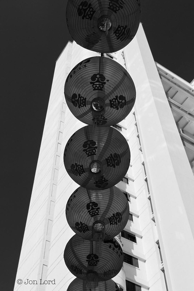 This is a high contrast abstract architectural black and white photo of a string of Chinese paper lanterns and a high-rise apartment block.
The camera is looking upwards at a near vertical angle. In the foreground is a string of six spherical Chinese paper lanterns strung from the middle of the lower margin up to the middle of the upper margin, they do actually slant a few degrees to the right as they stretch upwards. The lanterns are rendered dark grey, near black though they are very slightly transparent with the supporting metal wires visible within. On the base of these are four bold black Chinese characters. In the near background is a brilliant white, near luminous, high-rise apartment building with about twenty or more floors visible. The block is a near rectangular shape and is viewed from near the end of the building. Behind is a clear and cloudless sky rendered jet black.
The location: Chinatown, Singapore (2015).
