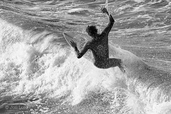 This is a black and white photo of a surfer riding along on the crest of a wave. 
In the centre of the image is a male surfer, with his back to the camera, on his surfboard riding along at right angles to the crest of a breaking wave and moving from right to left and away from the viewer. The wave stretches from the lower right corner to the upper left corner, with the surf on the left and sea on the right. Our surfer has dark hair, is wearing a black wetsuit and has his arms outstretched but with his hands raised upwards as if to aid his balance. The board is angled upwards and tilted to the left, towards the breaking surf. Surrounding the surfer is a thin cloud of water droplets lit by the still low morning sun.
The location: Hermosa Beach, California (2014).