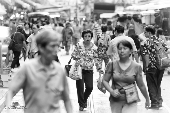 This is a black and white street photo of a crowded street in the Far East.
In the image's centre is a medium height, middle-aged Chinese lady, with a stern and determined expression, walking and facing towards the camera. She has severe permed dark hair, is wearing steel rimmed glasses, a white and floral blouse and black trousers. On both arms are plastic shopping bags and in her right hand she is holding a purse. At her feet is a steel rail track. Surrounding our lady is a crowd of lightly dressed people walking in both directions along the middle of the street. Almost all this crowd is heavily blurred out of focus. Either side of the street are the blurred outlines of market stalls.
The location: North Point, Hong Kong (2014).
