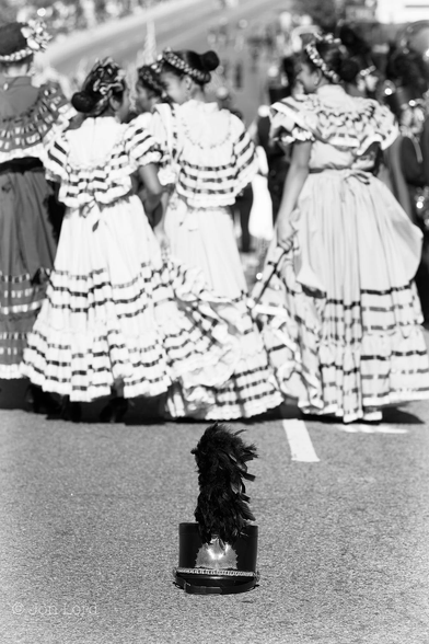 This is a black and white street photo of a parade with a lone black cap resting in the road.
Filling the lower third of the image is the surface of a road with a partially obscured white line visible to the lower right of centre. Below the centre and facing the camera is a single black cap with a large black feather rising from the top. The cap is cylindrical with a peak on the base, a silver badge on the front and a round flat top. About two metres beyond and slightly blurred out of focus, is a group of young, dark haired girls wearing long ornate dresses, they are standing shoulder to shoulder with their heads facing the girl on the left. The street slopes downwards and a crowd of people can be seen ahead, though heavily blurred.
The location: N Hill Street, Los Angeles. The occasion: The Chinese New Year parade (2014).