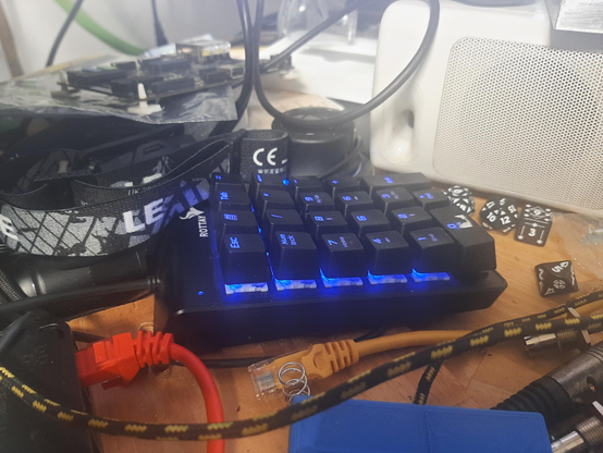 A cheap numeric keypad amidst a mess of ethernet cables, a 3d printed phone charge dock, 1990's speakers, a snickerdoodle black pysmasher board, multi-sided dice, a 3d spacemouce, a raspberry pi, an LED head lamp, etc. as is typical just to the left side of most people's keyboards.