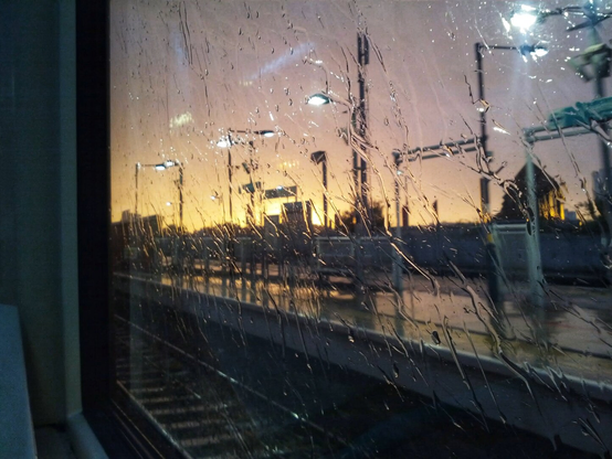 A moody view from inside a train, looking out through a rain-streaked window at a station platform during a late summer sunset. Streaks of water run vertically across the glass, distorting the scene. Beyond the wet window, the sky glows a warm orange and yellow, contrasting with the dark silhouettes of overhead gantries, lampposts, and railway tracks. The platform appears wet and reflective under the dim light, with distant buildings vaguely visible.