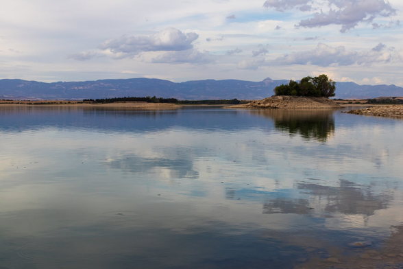 View of a calm lake reflecting the sky with white and gray clouds. On the right, there is a small rocky island with trees, and in the background, some mountains.