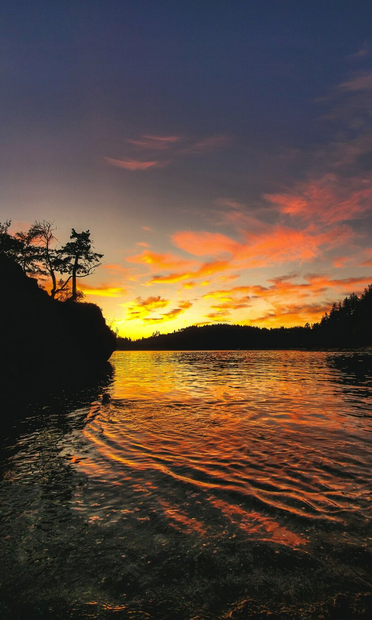 Vibrant sunset over a calm body of water, with fiery orange and pink clouds glowing against a darkening blue sky. The water reflects the warm colors in rippling patterns, while silhouetted trees and cliffs frame the scene on the left and right.