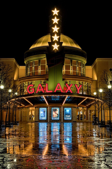 A color portrait photo of the front of a movie theater on a rainy night. The theater has a round entrance with a domed roof. There is a vertical lighted sign at the top with five five pointed white stars. There is are windows below the dome and below that is a marque that says in bright red light, "Galaxy." The rounded entrance is lit with small light bulbs. In the center are three movie poster that are backlit with blue lights but are too far away to read. The colors of all of the light are reflected on the wet concrete in front of the entrance.