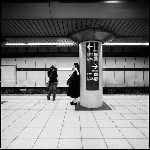 black and white photograph of two figures on a tokyo metro platform. a woman facing away from the camera, and a carmelite(?) nun in profile. arrows point in many directions... what can it all mean?