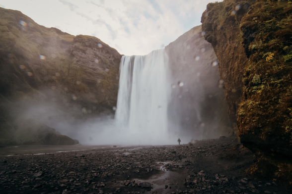 A wide, misty view of a powerful waterfall cascading from a high cliff into a rocky riverbed. Water droplets blur parts of the camera lens, adding a dreamy effect. A small lone figure stands near the base of the falls, dwarfed by the immense scale of the cliffs and rushing water. Moss-covered rock walls frame the scene, with cloudy sky above.