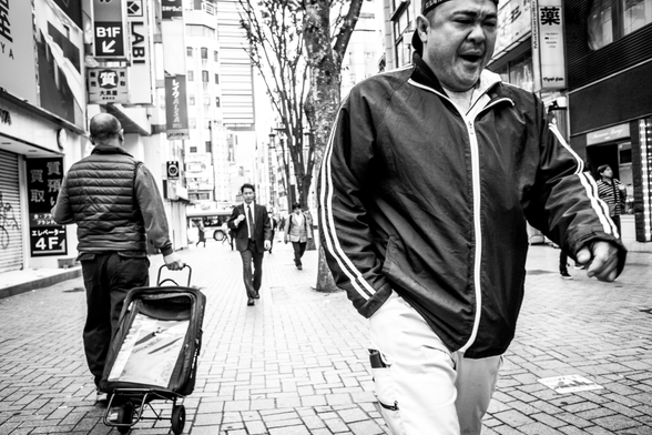Home que badalla mentre camina pels carrers de Tòquio, al darrere i té un salaryman i un altre home amb un carret.