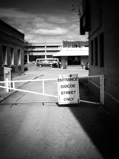 A white metal barrier blocks the entrance to a parking lot a white sign on the barrier reads "Entrance Simcoe Street Only". Buildings can be seen on both sides and in the background as well as some vehicles. Clouds also fill the sky in this black and white image.