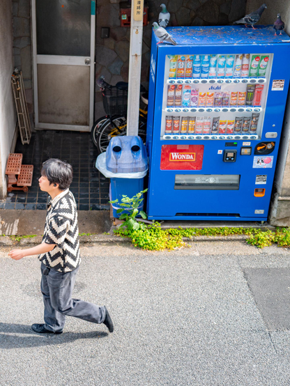 Ein Mann geht an einem blauen Getränkeautomaten in Tokio vorbei, während mehrere Tauben auf dem Automaten sitzen.