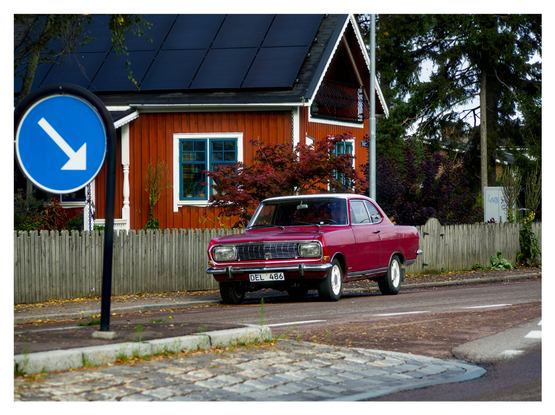 En liggande bild på en röd Opel Rekord Coupé med vitt tak. Den står parkerad vid trottoaren och ses snett framifrån.