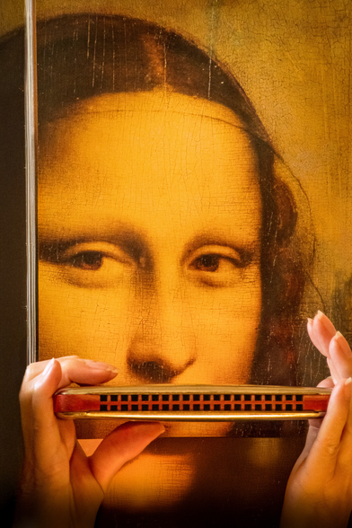 Das Bild zeigt eine Nahaufnahme eines Ausschnitts der Mona Lisa. Vor dem Bild wird eine Mundharmonika gehalten, die den unteren Teil des Gesichts der Mona Lisa verdeckt. Die Hände, die die Mundharmonika halten, sind im Vordergrund sichtbar.