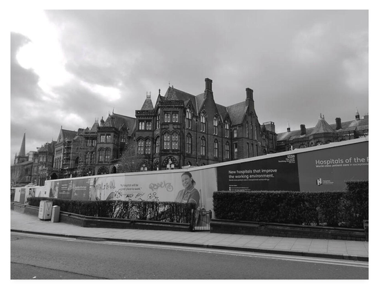 Black and white photo of an English Victorian hospital behind advertising hoardings.  