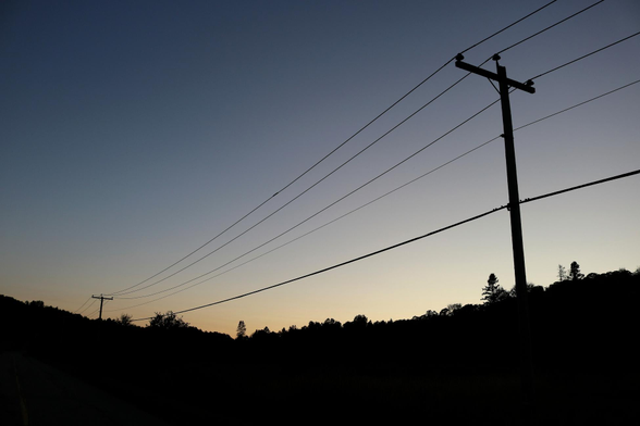Photograph of the side of a rural road at sunset, with a row of posts and power lines on the right side, and silhouettes of trees at the horizon. The sun has disappeared and the sky is blue at the top, and pale orange close to the horizon.