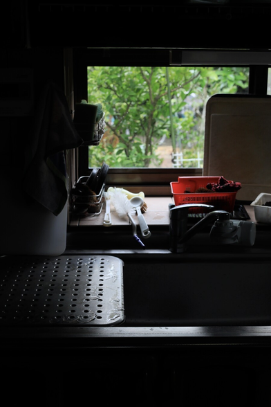 Sink by the small window.
Green plants in the yard are seen outside but it's dim inside.
kitchen tools put roughly are vaguely seen.