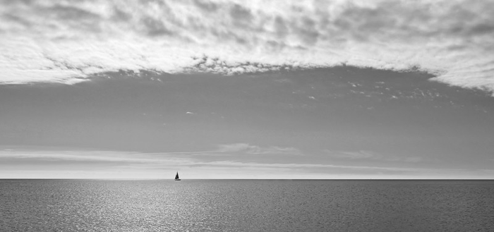 Man sieht eine ruhige See bis zum Horizon. Nur ein kleines Segelschif ist am Horizont zu erkennen. Im oberen Teil vom Foto sieht man einen Streifen von Wolken. Das Foto ist in Schwarzweiß.