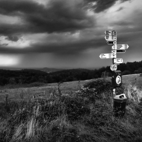 Ein Blick über kleine Berge mit einem dramatischen Himmel. In der Ferne kann man erahnen, dass es dort regnet. Am rechten Bildrand sind viele Hinweisschilder an einerm Metallpfosten angebracht, die die Entfernung zu den unterschiedlich Ausflugszielen anzeigen. Darunter ist ein Mülleimer. Das Foto ist in Schwarzweiß.
