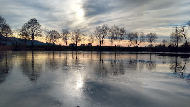 Eine Aufnahme von einem See im Winter. Die Wasseroberfläche ist fast zugefroren. Am anderen Ufer stehen Bäume ohne Blätter, die sich teilweise auf der Oberfläche vom See spiegeln. Am Himmel ist die Sonne durch leichte Schleierwolken zu sehen. Sie spiegelt sich auf der dünnen Eisschicht vom See. 