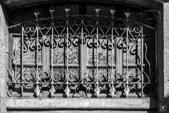 Black and white photo of a dusty paned window with an arched top set in a stone building with a decorative grate in front of it. There is small finger-created graffiti rubbed into the dust all over the window. 