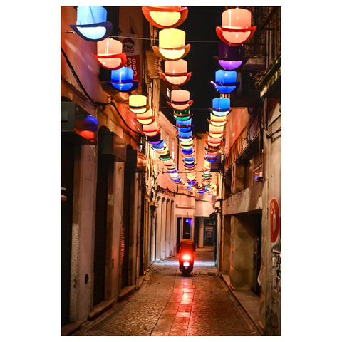 a narrow, cobblestone street at night, illuminated by a series of colorful, stacked hat-shaped lights hanging overhead. The lights are in various colors, including red, blue, green, and white,. The street is flanked by buildings with closed shutters and some signs. In the distance, a person on a scooter is driving away from the camera, with their taillight visible. (draft with mistral.ai, edited by author)