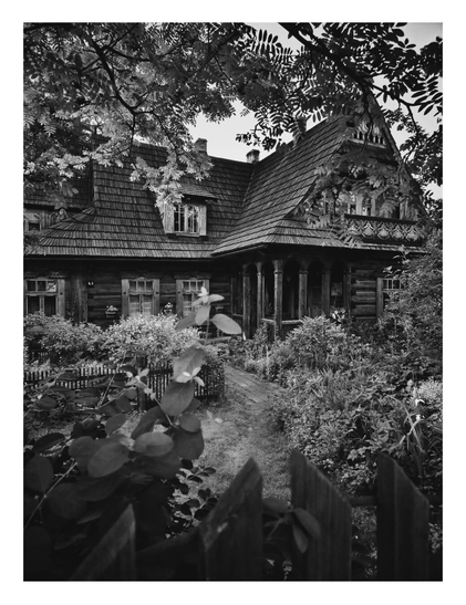 Black and white photo in portrait orientation of a traditional wooden house. The house has a multi-gabled, shingled roof and an ornate porch. A small path leads to the front door, and the house is surrounded by dense foliage and a wooden fence. Tree branches with leaves frame the top and foreground of the image.