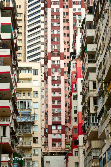 This is an abstract colour photo of a number of apartment buildings in a city setting, shot in portrait format. 
The camera is looking along a fairly narrow city street and raised upwards very slightly. Either side of the street are three medium-rise, closely spaced,  apartment blocks. On the left the closest building has long balconies running parallel to the street, each with a bright red stripe stretching along its base. Beyond is a cream coloured building that protrudes slightly and has balconies at right angles to the street below. This building has some minor construction work  being carried out with accompanying bamboo scaffolding covering one of the balconies. On the right side of the street the building are older with the colour being a weathered light brown. The facades are quite random, some having open balconies, some with those balconies enclosed with windows, some have external air conditioning units: some don't. At the far end of the street is an unseen 'T' junction with further apartment blocks facing the camera. These are much taller with twenty floors in view and none have balconies. The centre building is a light pink with the occasional thick maroon strip. Again randomly spaced A/C units and mismatched windows. To the left are more modern towers with an all uniform exterior. 
The location: Causeway Bay, Hong Kong (2013).

