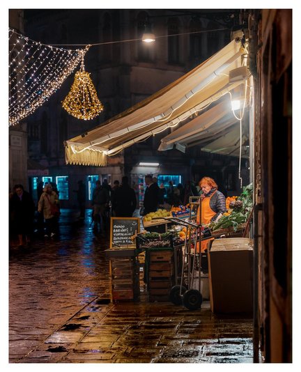 Eine Gemüseverkäuferin mit orangefarbener Schürze und Haaren steht draußen vor ihrem Laden. In der Umgebung sind Menschen und weihnachtliche Lichterketten die sich auf groben, nassen Strassensteine spiegeln zu sehen.