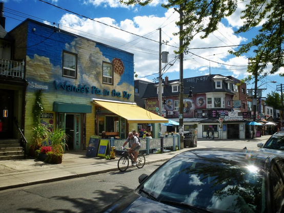 A bicyclist rides past a brightly painted building with clouds on it and large lettering reading "Wanda's Pie In The Sky". Cars are seen parked in the foreground and large planters counting flowers are on the sidewalk. A blue sky filled with white clouds and other buildings in the background are seen.