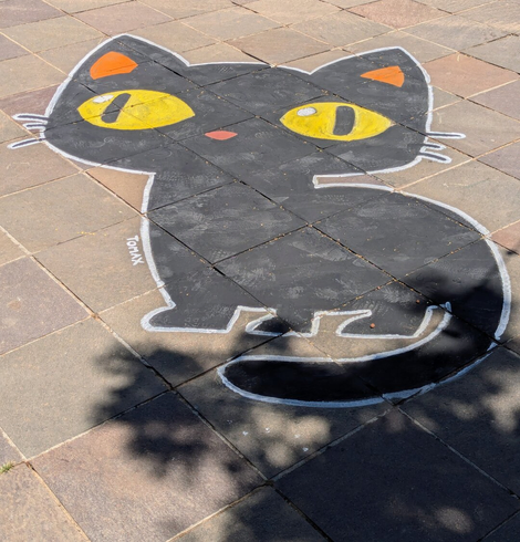 A top-down shot of a stylised black cat drawn in chalk on a paved outdoor surface. The cat has large, oval yellow eyes with black pupils, small orange inner ears, and a tiny red mouth. Its outline is drawn in white chalk, which also defines its whiskers and paws. The drawing extends across several grey and brown paving slabs, with some visible scuff marks on the black chalk. Shadows from an unseen object, likely a tree, fall across the bottom right of the image. A small, illegible signature, possibly "TOMAX", is written in white chalk near the cat's left front paw.