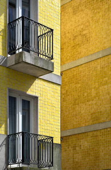 Fotografía en la que se ven parcialmente dos fachadas una detras de la otra, la de la derecha con dos balcones y baldosas amarillas, la de la derecha se ven baldosas amarillas separadas por franjas de hormigon, con una curiosa perspectiva dando a la imagen una sensación minimalista y abstracta.
