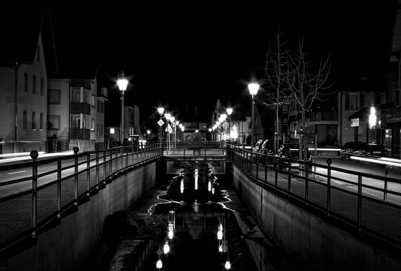 Foto der Hauptstrasse in meinem Ort bei Nacht. Diese wird durch einen kleinen Entlastungskanal geteilt. Die Strassenlaternen leuchten und spiegeln sich in dem wenigen Wasser im Kanal. Das Foto ist in Schwarzweiß.