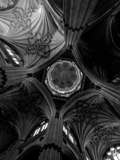 Una fotografía en blanco y negro del techo abovedado de una gran estructura arquitectónica, probablemente una catedral o iglesia, tomada desde abajo mirando hacia arriba. En el centro superior de la imagen, se aprecia una cúpula circular adornada con intrincados detalles radiales y una apertura superior que permite el paso de luz, creando un punto focal. Alrededor de esta cúpula, se extienden complejas bóvedas de crucería góticas, con nervaduras pronunciadas que convergen y forman elaborados patrones decorativos, algunos semejantes a rosetones florales o geométricos. Se distinguen pilares robustos que se elevan desde los bordes de la imagen para soportar estas bóvedas. La iluminación es dramática, con fuertes contrastes entre las áreas brillantes iluminadas por la luz que entra por las ventanas alargadas visibles en los laterales y la cúpula, y las zonas en sombra profunda, lo que realza la profundidad y la grandiosidad de la arquitectura.