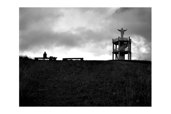 A black and white photo of a hillside under a dramatic cloudy sky. On the left, a lone person sits quietly on a bench, while on the right, a tall structure with a statue of Christ with outstretched arms rises above the landscape. A contemplative scene for #SilenceSunday