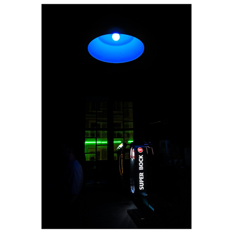 This image shows a dimly lit bar at night. The focal point is a "Super Bock" branded beer dispenser, illuminated by its own lights. Above the machine, a large circular blue light hangs from the ceiling, casting a glow downward. The background is dark, with some green neon lights visible through a window, adding to the bar's vibrant, nighttime atmosphere. (draft with mistral.ai, edited by author)