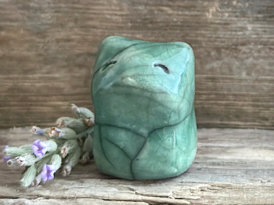 against a background of grey-brown driftwood, a very small raku ceramic fox sculpture glazed in a soft forest-green colour. it has a gentle, happy face and is turned slightly to the left. there's some lavender next to it on the left.