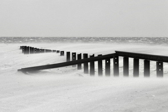 das bild zeigt eine schwarz-weiß-aufnahme eines strands bei stürmischem wetter. ein buhne aus stahlträgern verläuft von rechts nach links oben in richtung meer. auf den trägern liegen ebenso stahlträger. an einer stelle ist ein liegender träger heruntergefallen.
im hintergrund ist ein schmaler streifen meer zu sehen. der himmel darüber scheint wolkenfrei und sonnig. der sandstrand ist vom wind verweht, so dass es aussieht, als ob sich ein sandsturm über dem strand befindet.