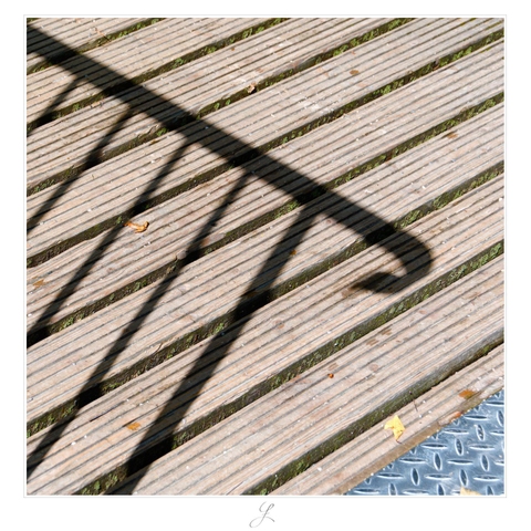 The square photo shows part of the deck of a wooden bridge. The light gray-brown wooden planks run diagonally from the bottom left to the top right. From the left, a metal railing casts its shadow on the wooden deck. The shadow of the handrail runs diagonally from top left to bottom right, until just before the right edge of the picture halfway up the photo. The shadows of the railing struts form a diamond pattern with the thin dark gaps between the wooden planks. In the lower right corner of the picture you can see a silver metal plate with an x-shaped surface profile. It creates a triangular image closure.

AI disclaimer: Using my work, its meta data, written or derived description to create media with or train AI based systems is prohibited.