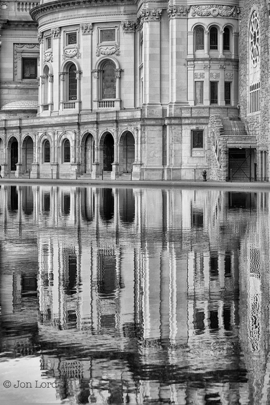 This is a black and white photo of an ornate building with its reflection in a ornamental lake.
The lower 60% of the image is filled with the dark waters of an ornamental city lake. On the far side at the edge of a lake is a grand, complex, marble like building with multiple arches, some containing steps up into the building. Above the arches is a short wall stretching across most of the facade that's in view. Above is a tall, semi-circular building's wall, again in marble or similar material. This wall has large rectangular windows with an arched top.Above the windows are some further but smaller rectangular windows. All of this is reflected into the dark waters below with just a few ripples disturbing the reflection.
The location: The christian science plaza, Boston (2013).