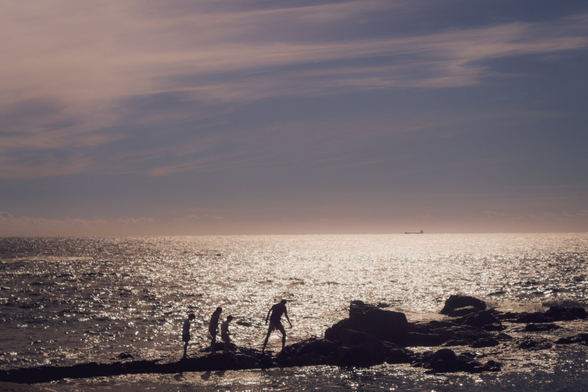 Silhouettes of four people walking on rocks by the sea at sunset, with the ocean shimmering under the sunlight and a ship visible in the distance.